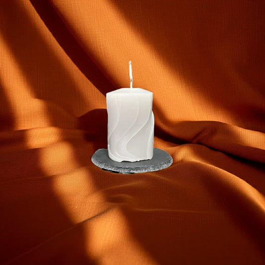 Weiße, handgemachte Kerze mit Wellenmuster auf dunklem Untersetzer vor orangefarbenem Stoff.