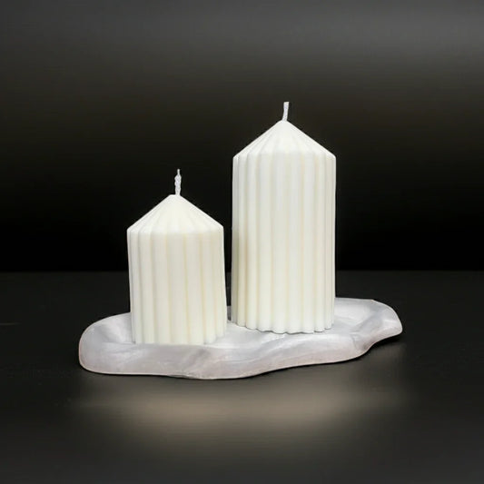 Zwei handgemachte, weiße Stumpenkerzen mit Rillen auf Marmorplatte, vor schwarzem Hintergrund.