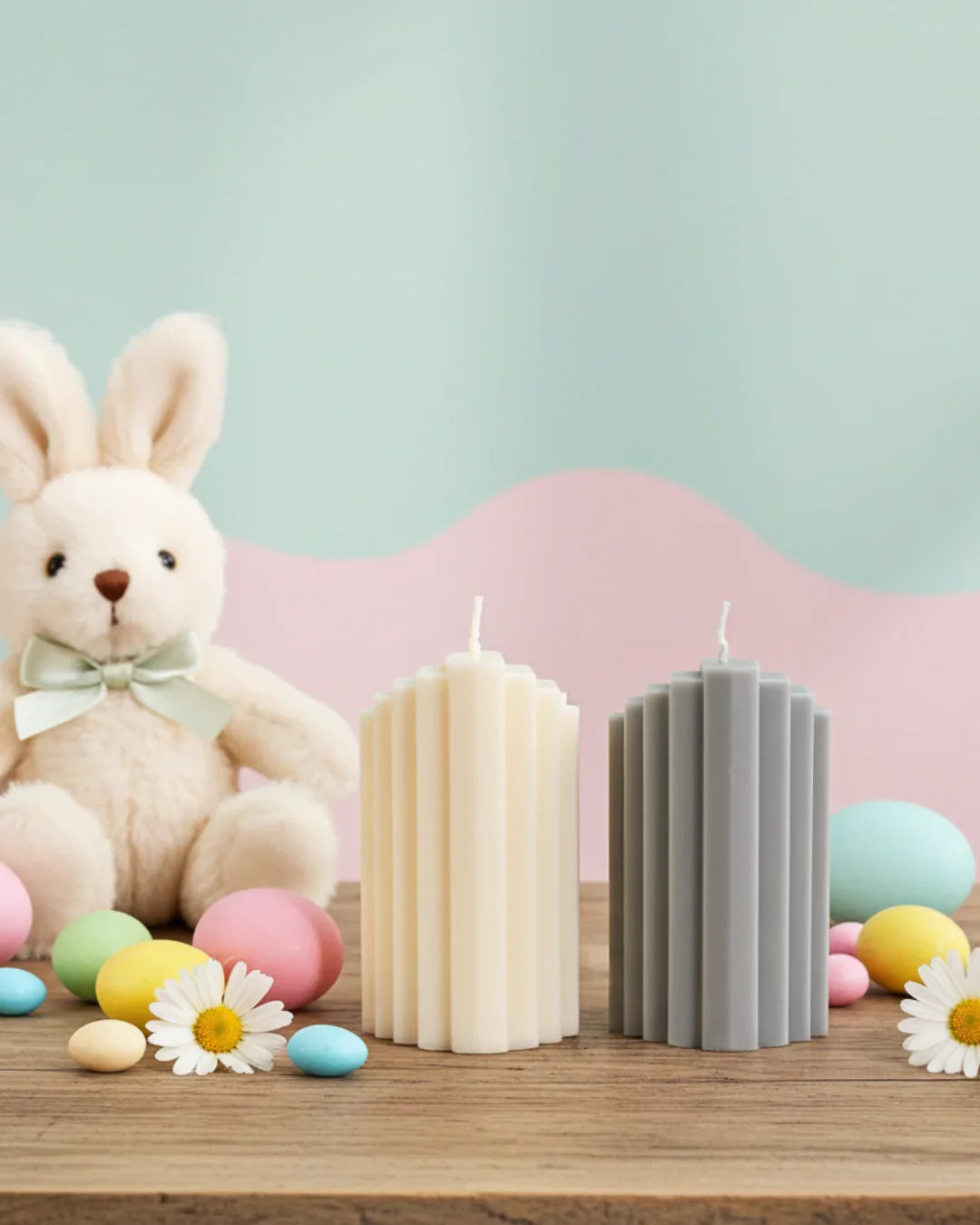 Zwei handgemachte geriffelte Kerzen, Osterdeko, Stoffhase und bunte Eier auf Holztisch