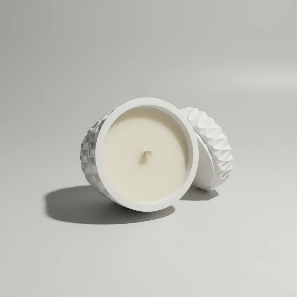 Weiße handgemachte Kerze in geometrischem Behälter mit Deckel auf hellem Hintergrund.