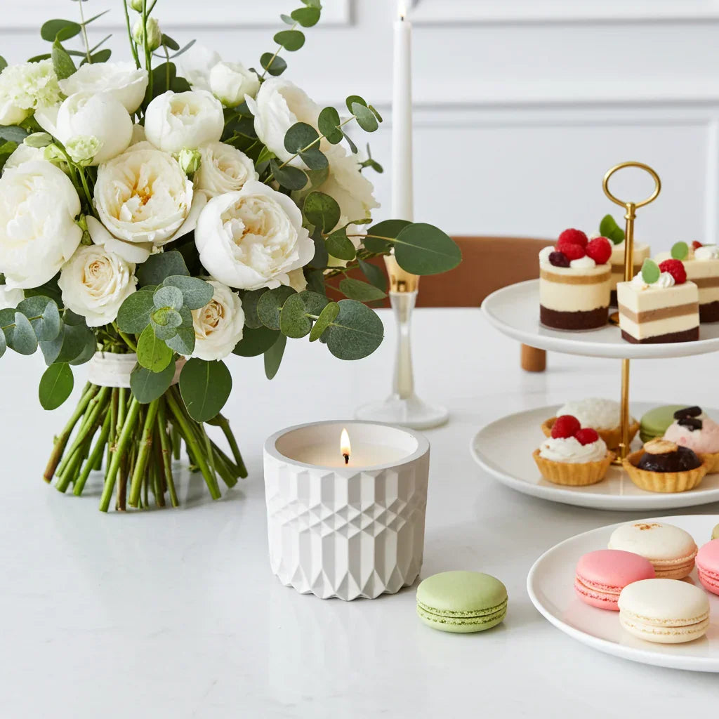 Handgemachte Duftkerze in weißem Gefäß, Blumenstrauß, Gebäck und Macarons auf Tisch