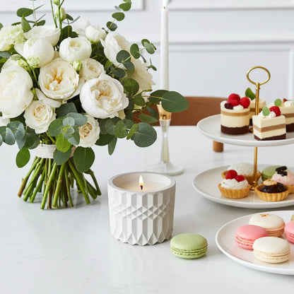 Handgemachte Duftkerze in weißem Gefäß, Blumenstrauß, Gebäck und Macarons auf Tisch