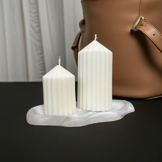 Zwei handgemachte, geriffelte Kerzen in Weiß auf einem marmorierten Kerzenteller, minimalistisch dekoriert.