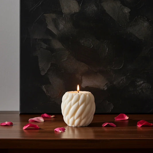 Handgemachte weiße Kerze mit Muster, brennend auf Holzoberfläche, umgeben von Rosenblättern