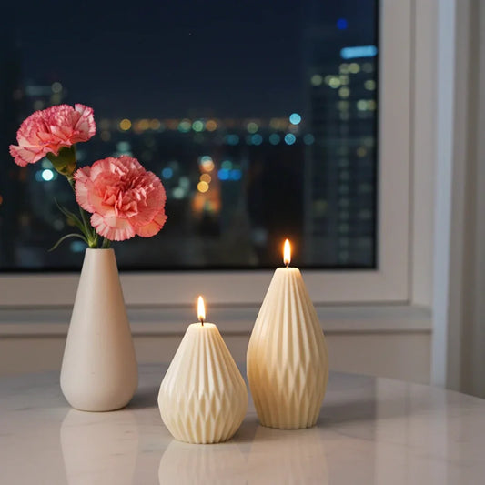 Zwei handgemachte, strukturierte Kerzen und Vase mit rosa Nelken auf Tisch am Fenster bei Nacht